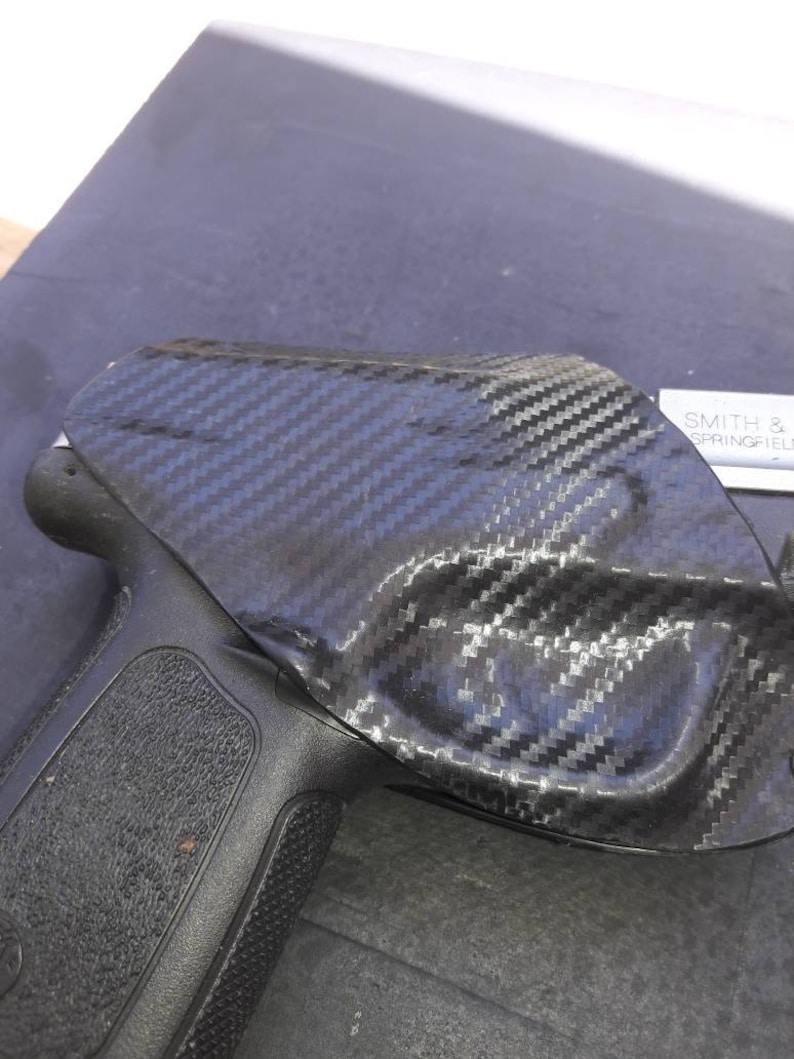 May include: A black handgun with a carbon fiber holster. The holster has a textured surface and is designed to fit snugly around the gun. The text "SMITH & WESSON" is visible on the holster.