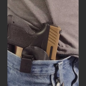 May include: A black and tan handgun with a black holster is clipped to a pair of blue jeans. A silver keychain with keys is attached to the jeans.