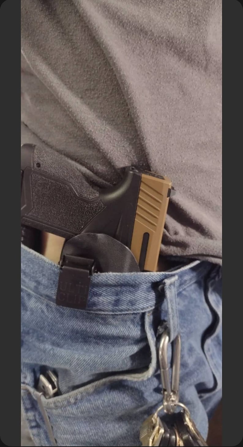 May include: A black and tan handgun with a black holster is clipped to a pair of blue jeans. A silver keychain with keys is attached to the jeans.