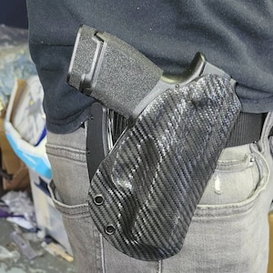 May include: A black handgun in a carbon fiber pattern holster, secured to a person's waist. The holster is attached to a black belt, and the person is wearing gray jeans and a black shirt. The handgun has a textured grip.