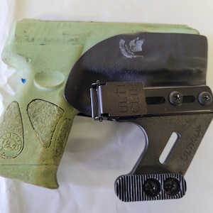 May include: A green handgun in a black holster with the text "ULTI CLIP" visible. The holster has a metal clip and is designed to be worn on the body. The handgun has a textured grip.