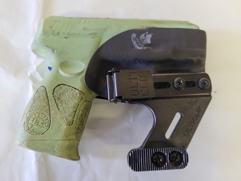 May include: A green handgun in a black holster with the text "ULTI CLIP" visible. The holster has a metal clip and is designed to be worn on the body. The handgun has a textured grip.