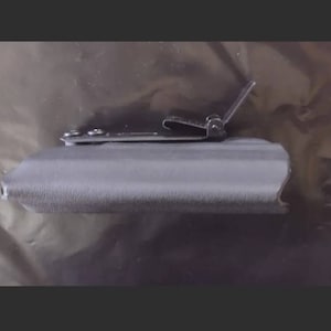 May include: A gray, textured holster with a metal clip. The clip is attached with small screws and is designed to secure the holster to a belt or other item. The holster appears to be made of a durable material.