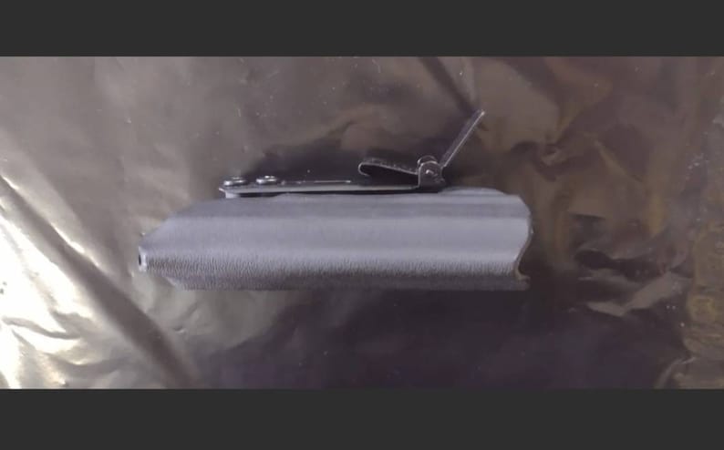 May include: A gray, textured holster with a metal clip. The clip is attached with small screws and is designed to secure the holster to a belt or other item. The holster appears to be made of a durable material.