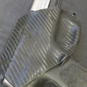 May include: A black carbon fiber holster with a silver handgun. The holster has a textured pattern and is designed to securely hold the firearm. The handgun is a Smith & Wesson, Springfield, MA USA.