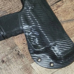 May include: Black carbon fiber holster with a textured weave pattern. The holster has two metal eyelets and is designed to hold a black handgun. The image is set against a wooden background.