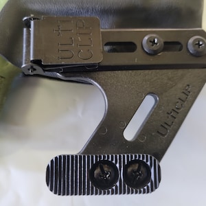 May include: A black UltiClip holster clip with the text "ULTI CLIP" visible. The clip is attached to a black holster and secured with screws. The clip has a textured, grooved base.