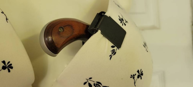 May include: A small, silver handgun with a wooden grip is tucked into a white fabric bra with a floral pattern. A black clip is attached to the bra.
