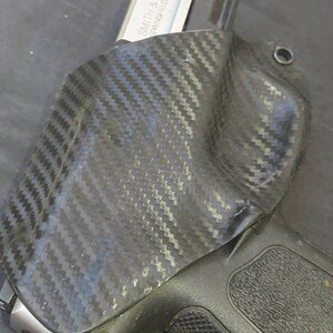 May include: A black carbon fiber holster with a silver handgun inside. The handgun has the text "SMITH & WESSON SPRINGFIELD, MA USA" on the side.