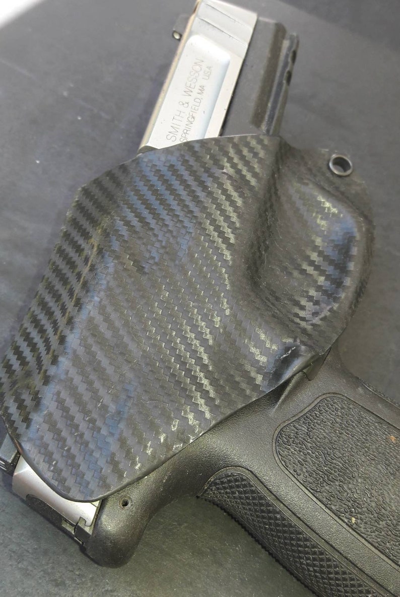 May include: A black carbon fiber holster with a silver handgun inside. The handgun has the text "SMITH & WESSON SPRINGFIELD, MA USA" on the side.
