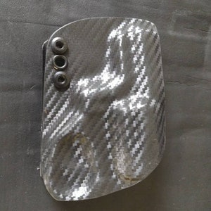 Puede incluir: Una funda negra de fibra de carbono con tres remaches negros. La funda tiene un patr&oacute;n texturizado y tejido y una forma curva. El art&iacute;culo est&aacute; dise&ntilde;ado para sujetar un objeto peque&ntilde;o.