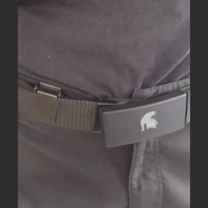 May include: A black belt with a silver buckle and a rectangular clasp featuring a silver Spartan helmet design. The belt is worn with dark gray pants.