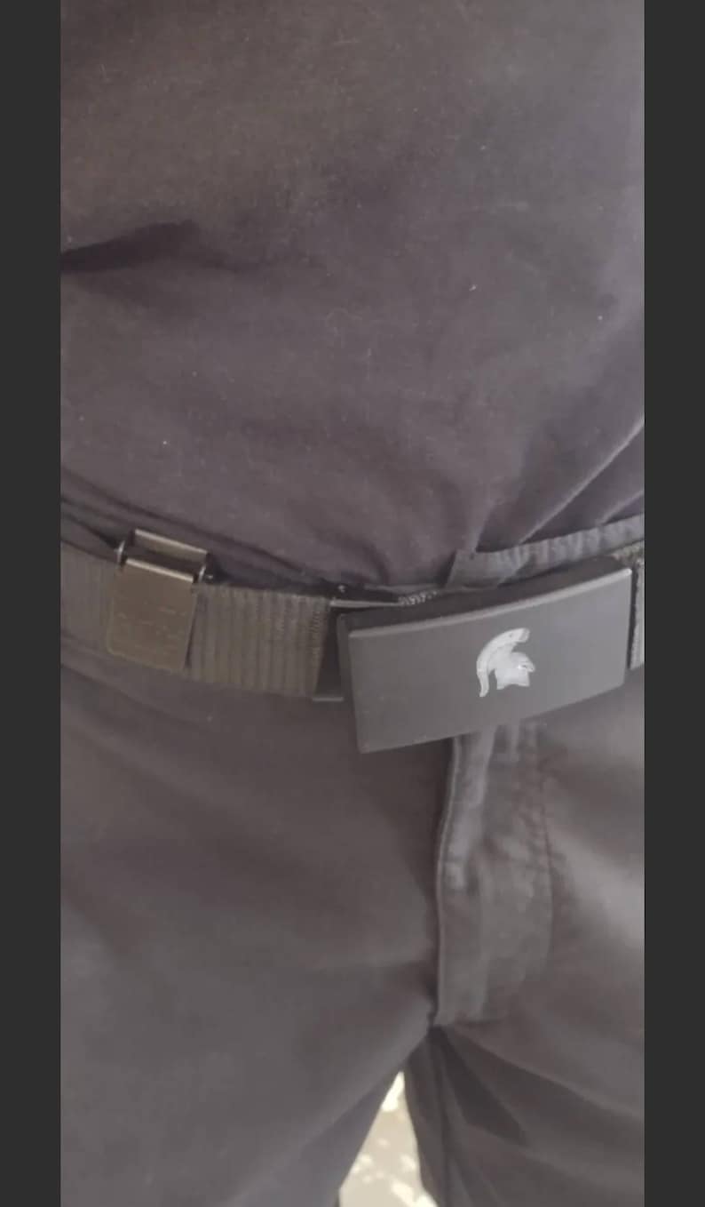 May include: A black belt with a silver buckle and a rectangular clasp featuring a silver Spartan helmet design. The belt is worn with dark gray pants.