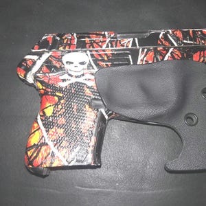 May include: A handgun with a red, orange, and black camouflage pattern and a white skull graphic. A black holster is placed next to the handgun.
