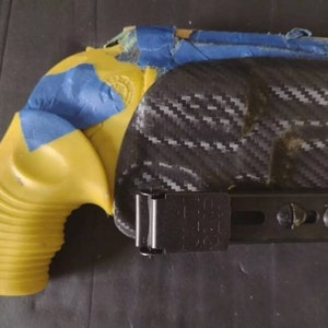 Puede incluir: Una pistola amarilla con cinta azul y una funda negra. La funda tiene un patr&oacute;n de fibra de carbono. La empu&ntilde;adura de la pistola es acanalada. La funda tiene un clip negro.