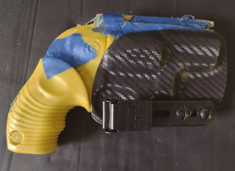 Puede incluir: Una pistola amarilla con cinta azul y una funda negra. La funda tiene un patr&oacute;n de fibra de carbono. La empu&ntilde;adura de la pistola es acanalada. La funda tiene un clip negro.