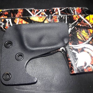 May include: A black leather holster with four metal eyelets for a firearm with a camouflage pattern. The firearm has a white Spartan helmet logo on the side.