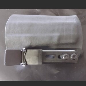 May include: A gray holster with a metal clip and screws. The holster is designed to hold an item, and the metal clip is used to secure it. The item is likely a tool or a small device.