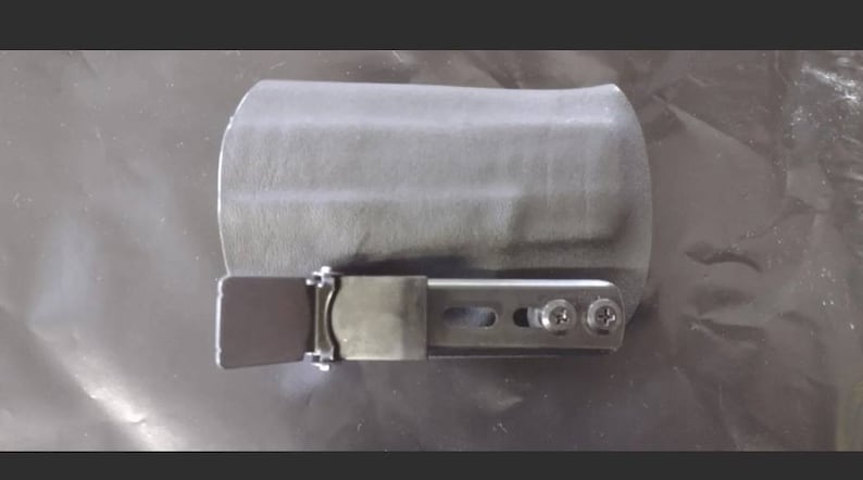 May include: A gray holster with a metal clip and screws. The holster is designed to hold an item, and the metal clip is used to secure it. The item is likely a tool or a small device.