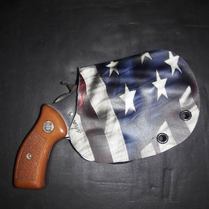 May include: A silver revolver with a brown wooden handle is in a leather holster with an American flag design.