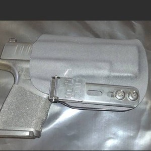 May include: A black handgun in a black holster. The holster has a metal clip and is attached to a metal bracket with three screws. The gun is partially visible, showing the grip and slide.