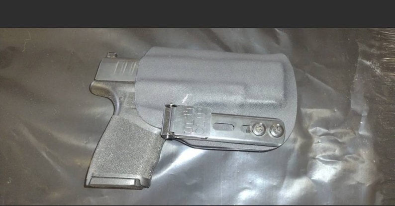 May include: A black handgun in a black holster. The holster has a metal clip and is attached to a metal bracket with three screws. The gun is partially visible, showing the grip and slide.