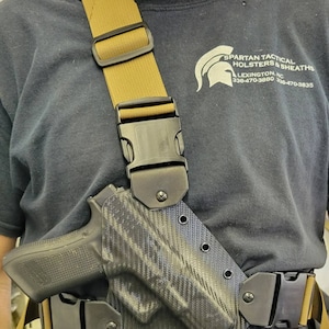 May include: A black and tan shoulder holster with a black handgun and a carbon fiber-patterned holster. The holster is attached to a tan strap with a black buckle. The strap is worn over a black t-shirt with the text "Spartan Tactical Holsters & Sheaths Lexington, NC 336-470-3880 336-470-3835".