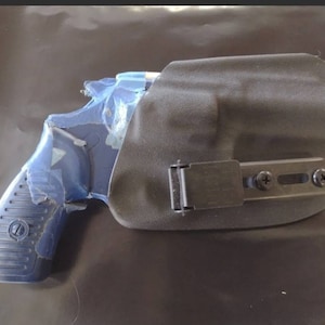 May include: A blue handgun with a black holster. The handgun is partially covered in blue protective material. The holster is black and has a metal clip and screws.