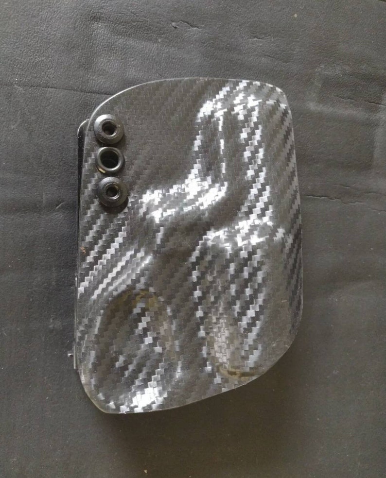 K&ouml;nnte beinhalten: Ein schwarzes und graues Holster aus Kohlefaser mit drei schwarzen Schrauben.