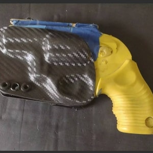 K&ouml;nnte beinhalten: Eine gelbe Pistole mit einem schwarzen Kohlefaserholster. Die Pistole hat einen strukturierten Griff und eine schwarze Sicherung.