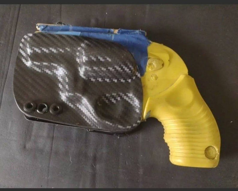 K&ouml;nnte beinhalten: Eine gelbe Pistole mit einem schwarzen Kohlefaserholster. Die Pistole hat einen strukturierten Griff und eine schwarze Sicherung.