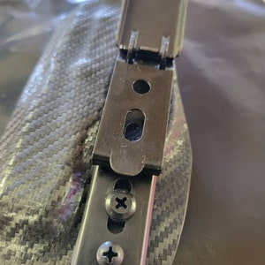 May include: A black metal clip with two screws, attached to a carbon fiber patterned surface. The clip has a hinged design and a rectangular shape with a small oval opening. The screws are silver with a cross-shaped indentation.