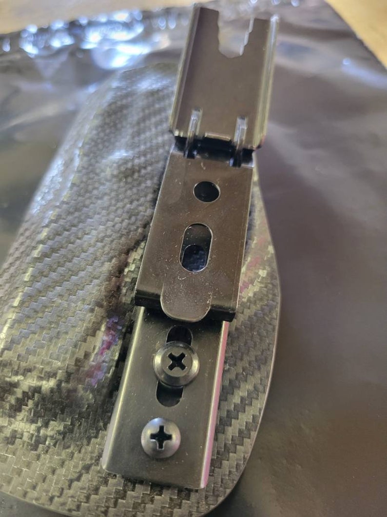 May include: A black metal clip with two screws, attached to a carbon fiber patterned surface. The clip has a hinged design and a rectangular shape with a small oval opening. The screws are silver with a cross-shaped indentation.