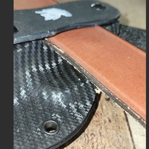 May include: Close-up of a leather strop with a black handle and a carbon fiber sheath. The strop is brown and the handle has two holes. The sheath has a woven pattern.
