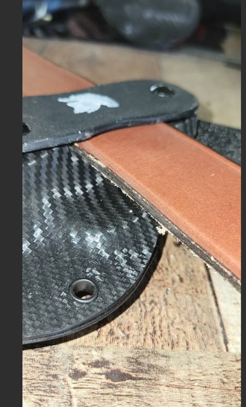 May include: Close-up of a leather strop with a black handle and a carbon fiber sheath. The strop is brown and the handle has two holes. The sheath has a woven pattern.