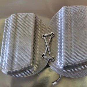 May include: A pair of black carbon fiber holsters, connected by a black cord laced through metal eyelets. The holsters have a textured, woven pattern. The overall shape is curved, with a rounded bottom.