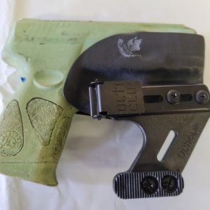 May include: A green handgun in a black holster with the text "ULTI CLIP" visible. The holster has a metal clip and is designed to be worn on the body. The handgun has a textured grip.