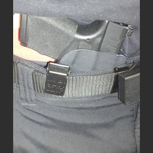 May include: Black handgun holstered inside the waistband, secured by a black belt with a metal clip. The holster is partially visible, and the gun's grip is exposed. The belt has a rectangular clip with the text "CLIP" on it.