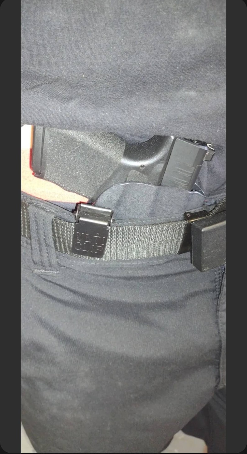 May include: Black handgun holstered inside the waistband, secured by a black belt with a metal clip. The holster is partially visible, and the gun's grip is exposed. The belt has a rectangular clip with the text "CLIP" on it.