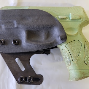 May include: A black holster with a textured surface is attached to a green handgun. The holster is secured with black screws and has a metal bracket. The handgun has a textured grip.