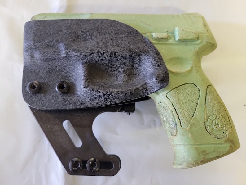May include: A black holster with a textured surface is attached to a green handgun. The holster is secured with black screws and has a metal bracket. The handgun has a textured grip.