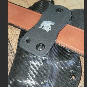 May include: A black holster with a carbon fiber pattern, featuring a brown leather belt and a black strap with a silver emblem. The holster is secured with screws.