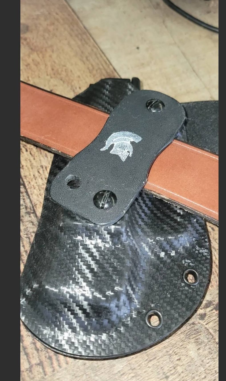 May include: A black holster with a carbon fiber pattern, featuring a brown leather belt and a black strap with a silver emblem. The holster is secured with screws.