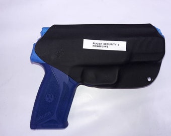 Könnte beinhalten: Ein schwarzes Lederholster mit einer blauen Ruger Security 9 Pistole darin. Das Holster trägt ein Etikett mit der Aufschrift "RUGER SECURITY 9 ROWB/LIWB".