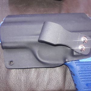 K&ouml;nnte beinhalten: Ein schwarzes Lederholster mit einer blauen und schwarzen Pistole. Das Holster hat einen Metallclip und zwei silberne Schrauben. Die Pistole hat den Schriftzug "RUGER" auf der Seite.