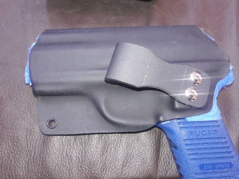 K&ouml;nnte beinhalten: Ein schwarzes Lederholster mit einer blauen und schwarzen Pistole. Das Holster hat einen Metallclip und zwei silberne Schrauben. Die Pistole hat den Schriftzug "RUGER" auf der Seite.