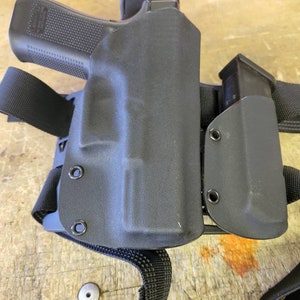 May include: A black tactical holster with a magazine pouch attached. The holster is made of a durable material and has a secure fit. The holster is designed to be worn on the hip or thigh.