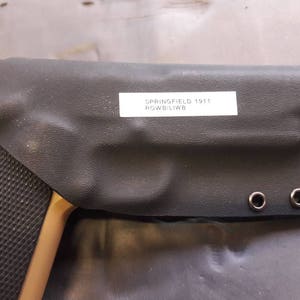 May include: A black leather holster with a label that reads "Springfield 1911 Rowblib". The holster has four black eyelets and a brown and black handgun is partially visible.