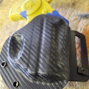 Puede incluir: Una funda de fibra de carbono negra con un clip para cintur&oacute;n. La funda est&aacute; dise&ntilde;ada para sostener una peque&ntilde;a pistola amarilla con una estrella en el lateral.