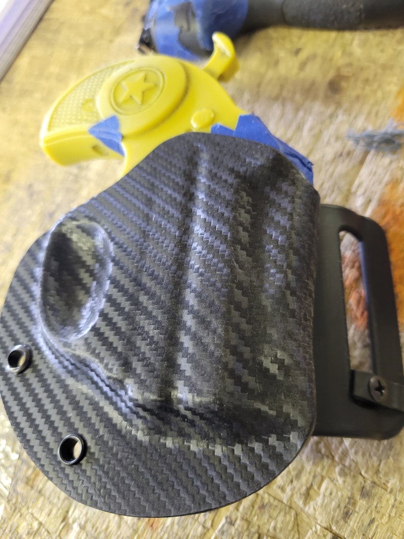 Puede incluir: Una funda de fibra de carbono negra con un clip para cintur&oacute;n. La funda est&aacute; dise&ntilde;ada para sostener una peque&ntilde;a pistola amarilla con una estrella en el lateral.